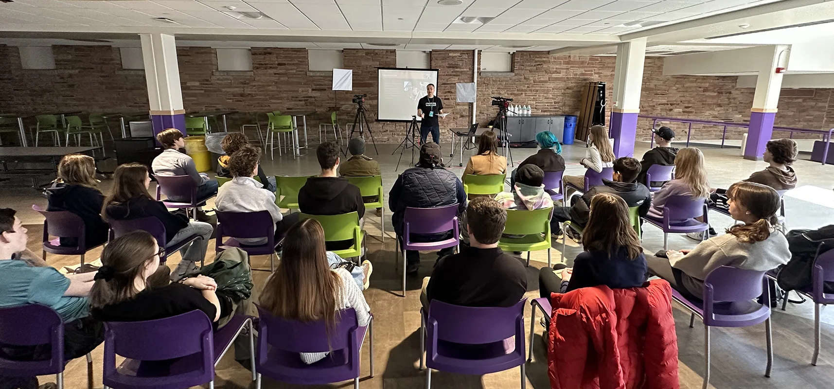 High school students seated in rows watching an instructor beside a projection screen and cameras on tripods at the Intro to PA workshop