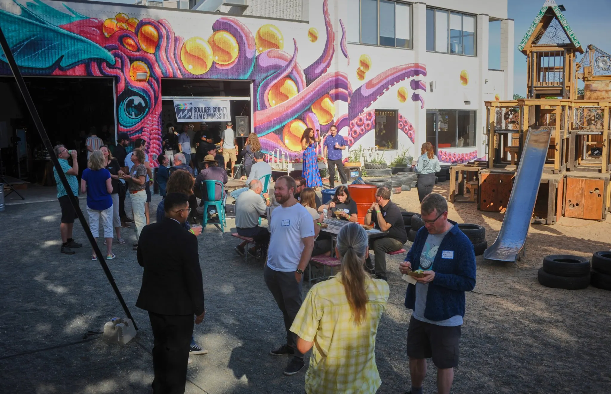 Crowd gathered outdoors at Junkyard Social Club in front of a colorful mural and the Boulder County Film Commission banner at the launch event