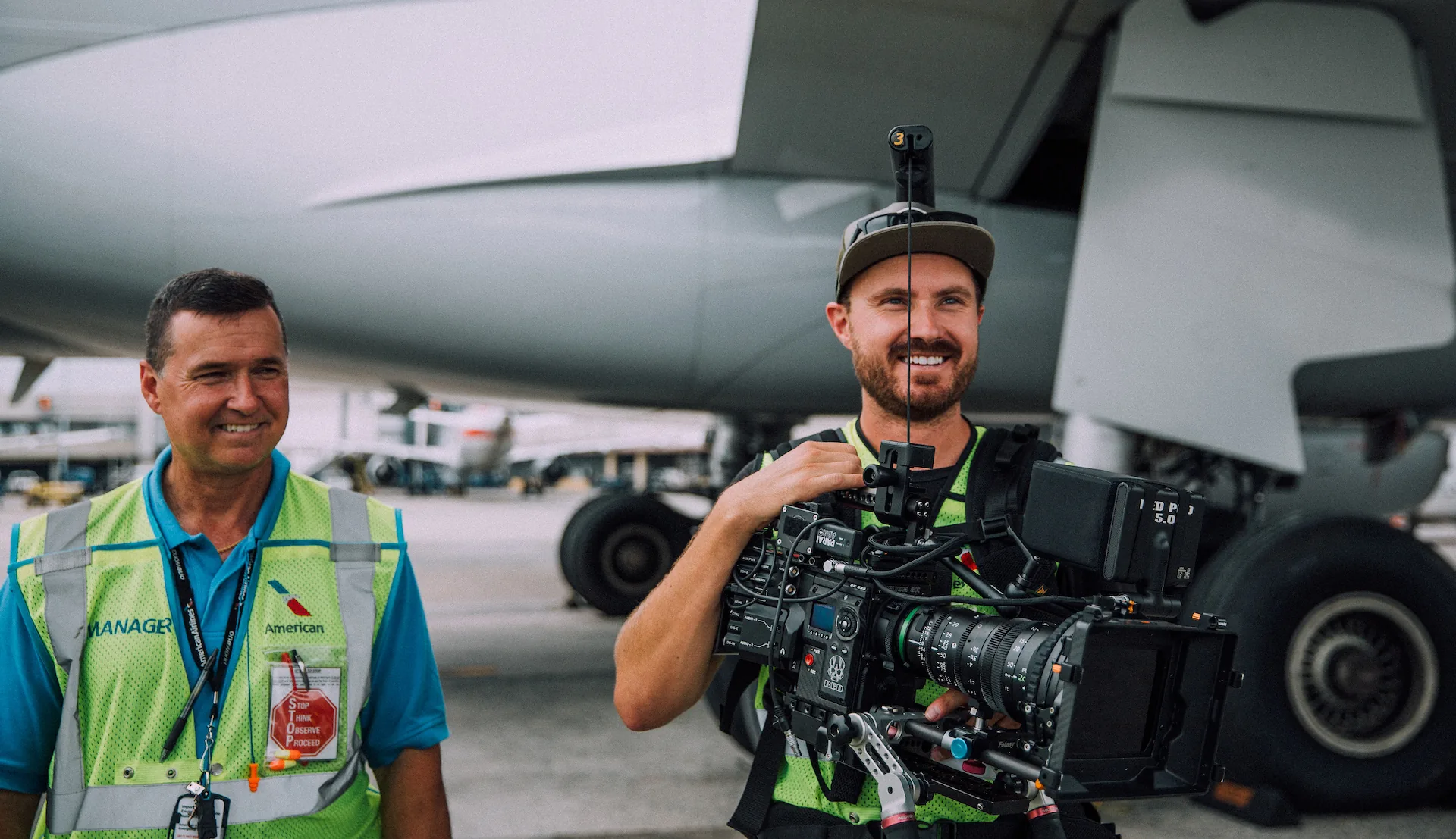 Camera operator with a cinema camera rig beside an airport ground crew member under an aircraft