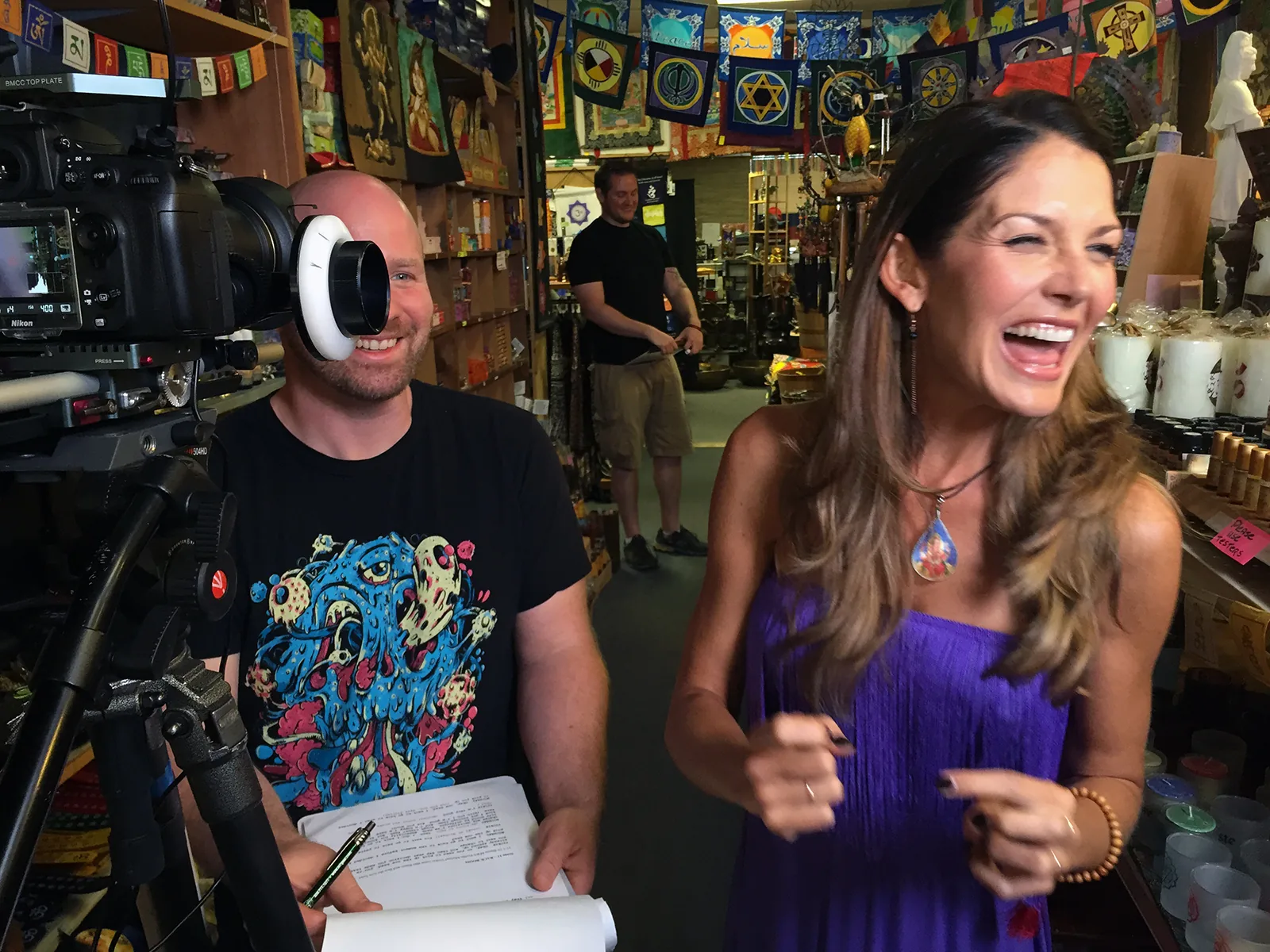 Laughing host in a purple dress being filmed in a colorful gift shop, with a smiling crew member holding a script beside the camera