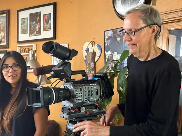 Camera operator setting up a cinema camera beside a smiling young woman in a room hung with framed pictures