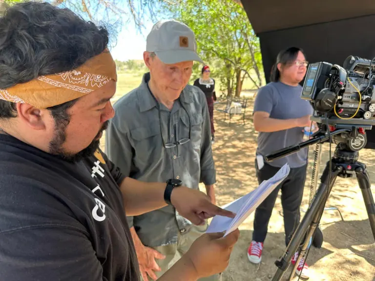 Documentary crew reviewing a script outdoors beside a cinema camera on a tripod