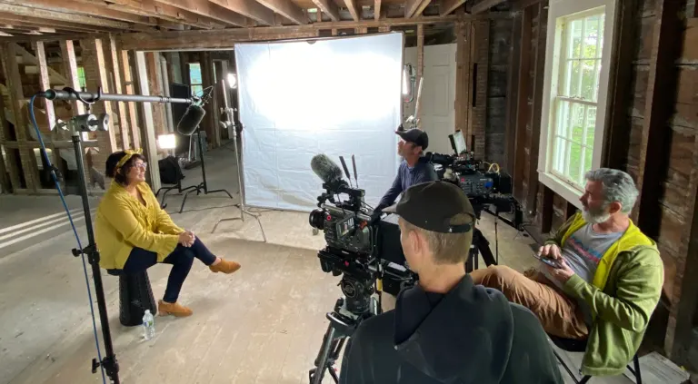 Film crew recording an interview with a seated woman against a white backdrop in a rustic wood-framed room