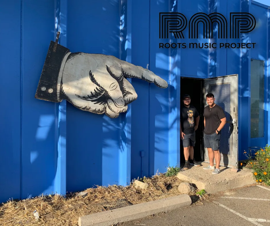 Large pointing-hand sign on the blue exterior wall of the Roots Music Project, with two men standing in the doorway