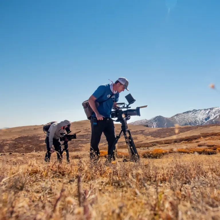 Two camera operators filming on a dry grassy plain with snow-capped mountains in the distance