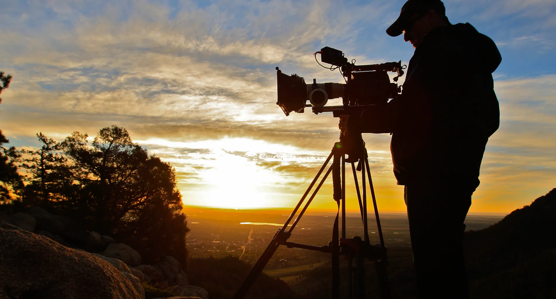 Filmmaker silhouetted against a sunset, operating a cinema camera on a tripod at a rocky overlook