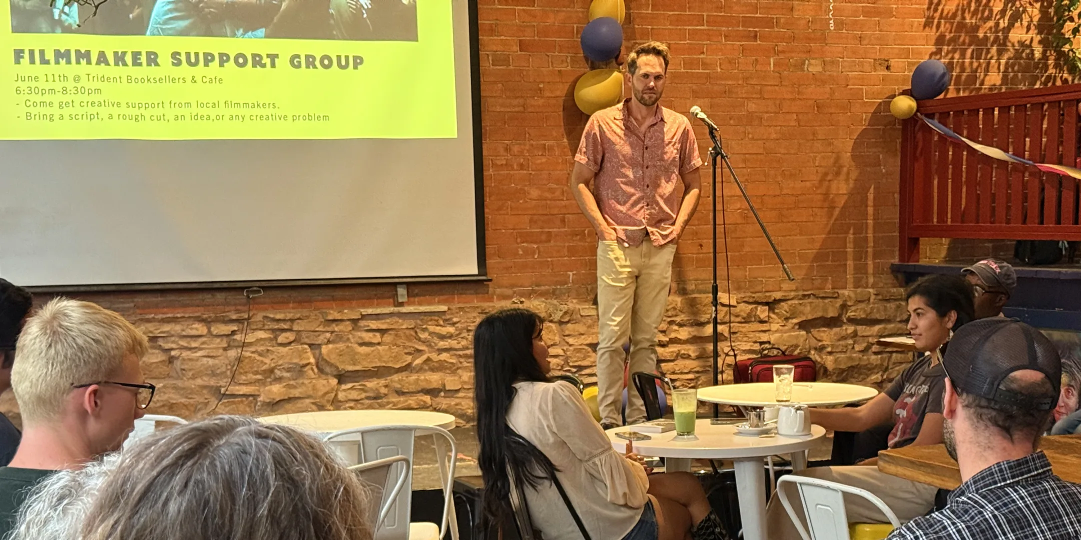 A filmmaker speaking to a small audience on the patio at Trident Booksellers and Cafe, beside a screen announcing the Filmmaker Support Group
