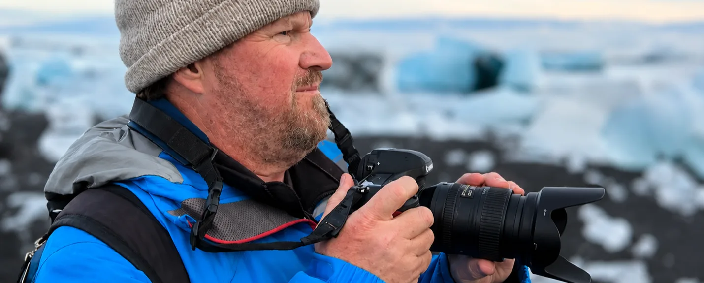A photographer in a blue jacket and knit cap framing a shot with a DSLR camera beside a glacial lagoon
