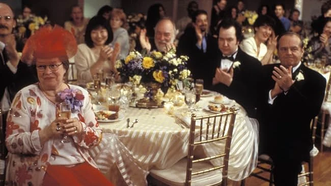 Warren Schmidt in a tuxedo and other guests applaud at a wedding reception table as a woman in a red feathered hat holds a champagne glass.