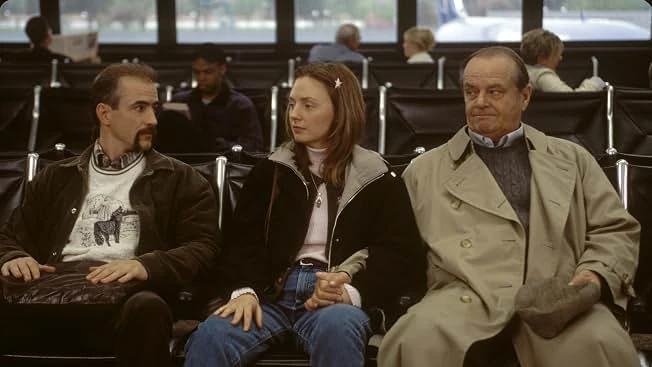 Warren Schmidt in a trench coat sits in an airport gate lounge beside a young woman and a man with a mustache.