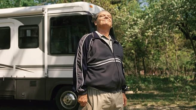 Warren Schmidt, in a track jacket with a cap in hand, turns his face to the sun beside a motor home among trees.