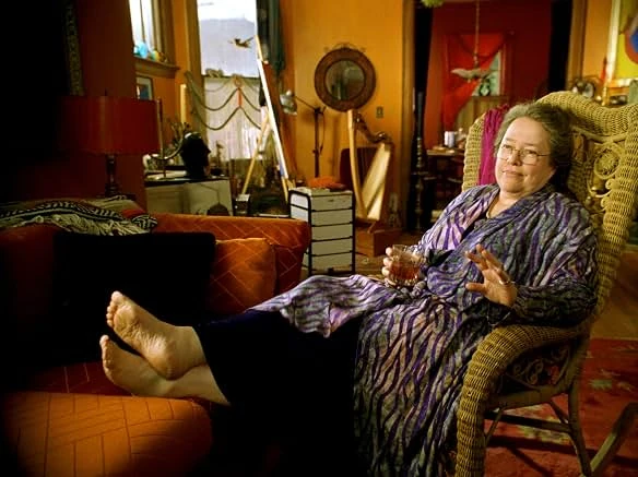 A woman in a purple striped robe relaxes in a wicker chair with her bare feet up and a drink in her hand.