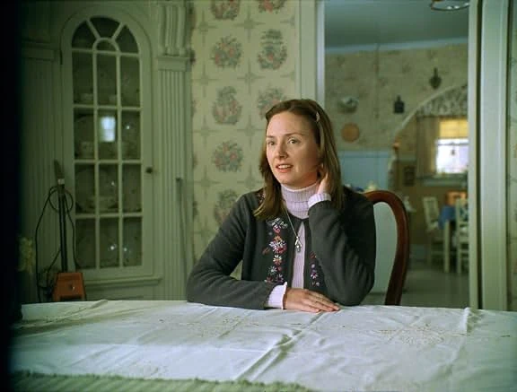 A young woman in a cardigan sits at a dining table with a white cloth in a house with floral wallpaper.