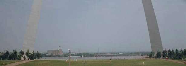 The legs of the Gateway Arch frame a grassy lawn with scattered people and a wide river under a hazy sky