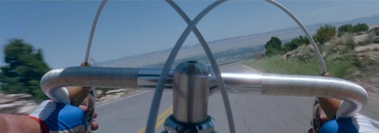 A rider's view over chrome handlebars and gloved hands, descending a winding high desert road under a blue sky