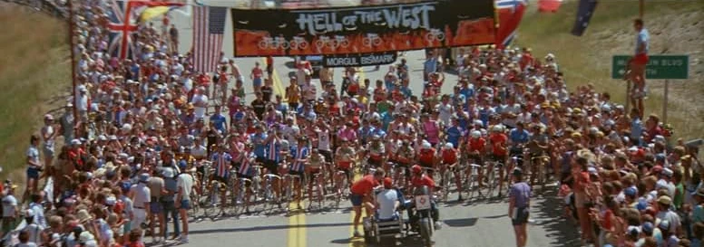 Cyclists pack the start line under a Hell of the West banner on a road lined with spectators and flags