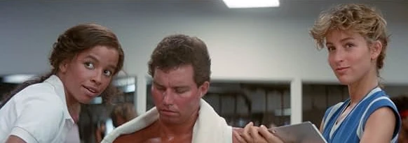 Two young women flank a shirtless man with a towel over his shoulders, one holding a clipboard
