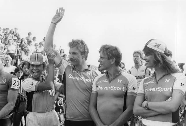 A mustached cyclist in a ShaverSport jersey raises his arm beside teammates as a crowd looks on, in black and white