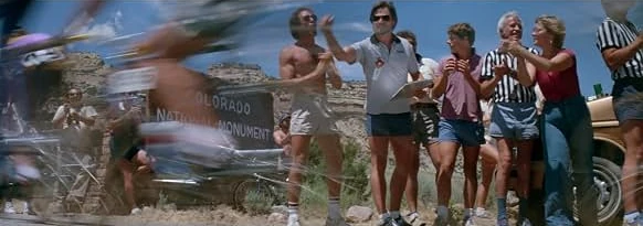 Spectators cheer beside a Colorado National Monument sign as racing cyclists blur past in the foreground