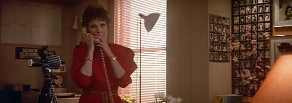 A woman in a red dress talks on the phone in an office with a camera on a tripod and a wall of headshots