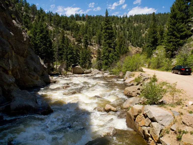 A whitewater creek rushing over boulders beside a dirt road and a parked car, with a rock cliff and conifer forest under blue sky.