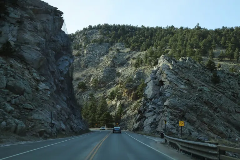 A two-lane highway curving between steep rock walls and pine-dotted slopes, with two cars ahead and a guardrail on the right.