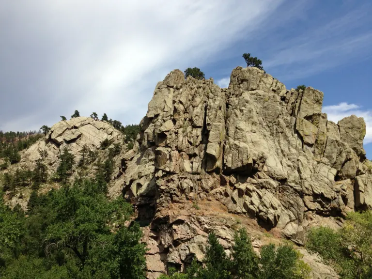 A tall fractured rock buttress topped with pines rising above green trees, beside a rounded rock dome under a streaked blue sky.