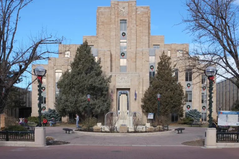 Symmetrical sandstone courthouse with a central clock tower, seen across a brick mall and a plaza with a fountain under a blue winter sky.