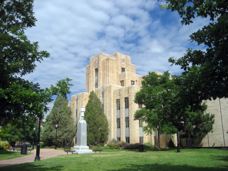 Tan sandstone courthouse with a stepped clock tower behind a soldier monument, evergreens and a green lawn under a partly cloudy sky.