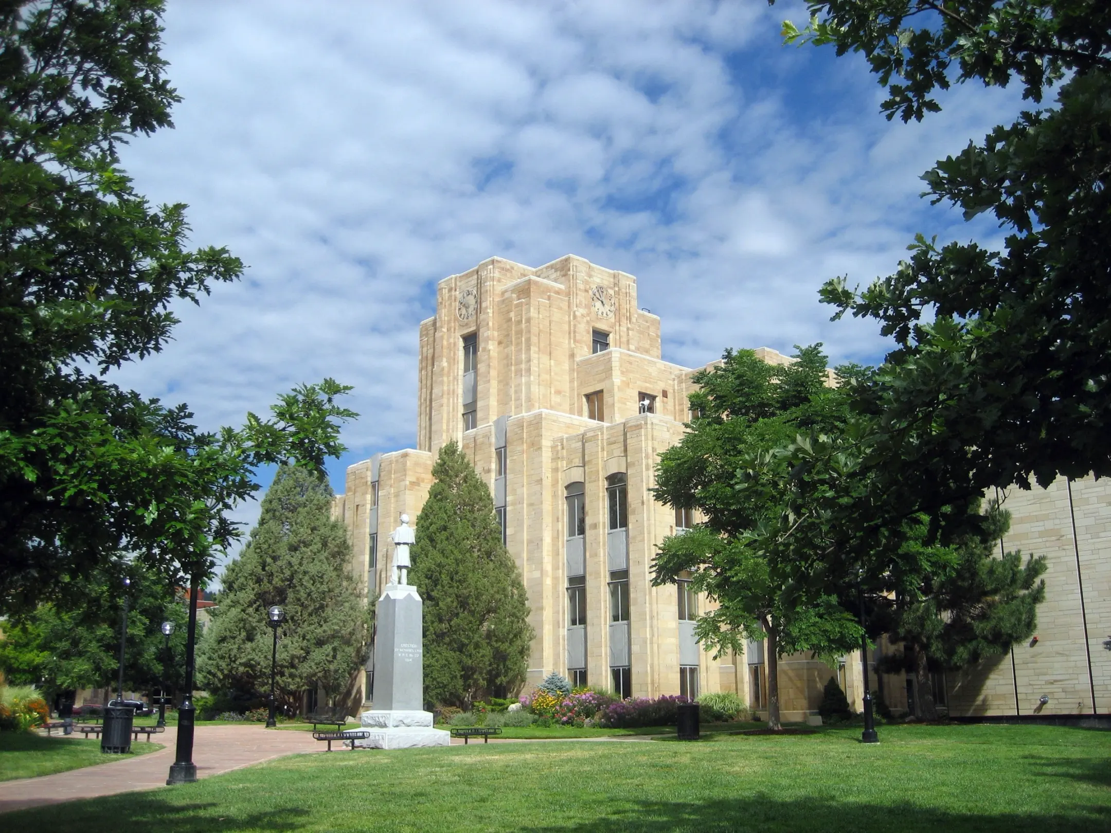 Tan sandstone courthouse with a stepped clock tower behind a soldier monument, evergreens and a green lawn under a partly cloudy sky.