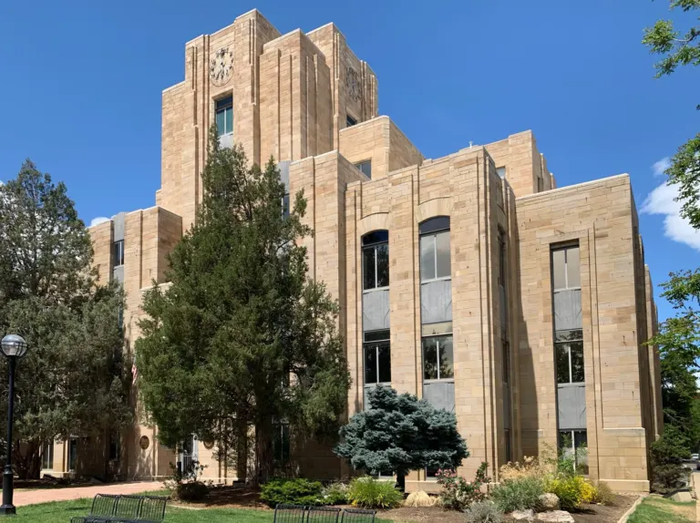 Corner view of a stepped sandstone courthouse tower with clock faces, tall window bays and evergreens under a clear blue sky.