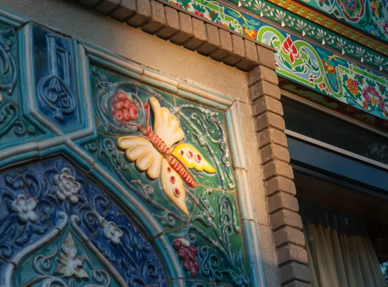 Glazed ceramic relief of a cream and yellow butterfly among green and blue scrollwork, below a brightly painted cornice.