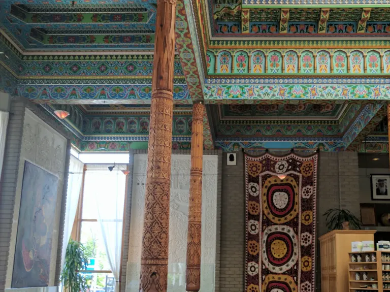 Carved cedar columns rise to a brightly painted coffered ceiling, beside white carved plaster panels and a hanging embroidered textile.