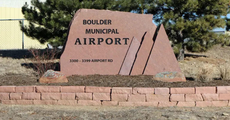 Red sandstone sign reading Boulder Municipal Airport, set behind a low stone wall in front of pine trees.