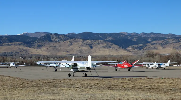 Small propeller planes tied down on an airport ramp, with dry grass in front and tan, pine-covered foothills under a clear blue sky.