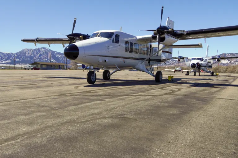 White twin-engine propeller plane parked on an asphalt ramp, with a low gray building and the snow-dusted Flatirons behind.