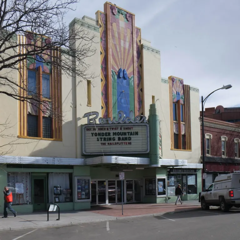 Cream stucco Art Deco theater front with tall pastel terracotta sunburst panels above a Boulder marquee and silver canopy.