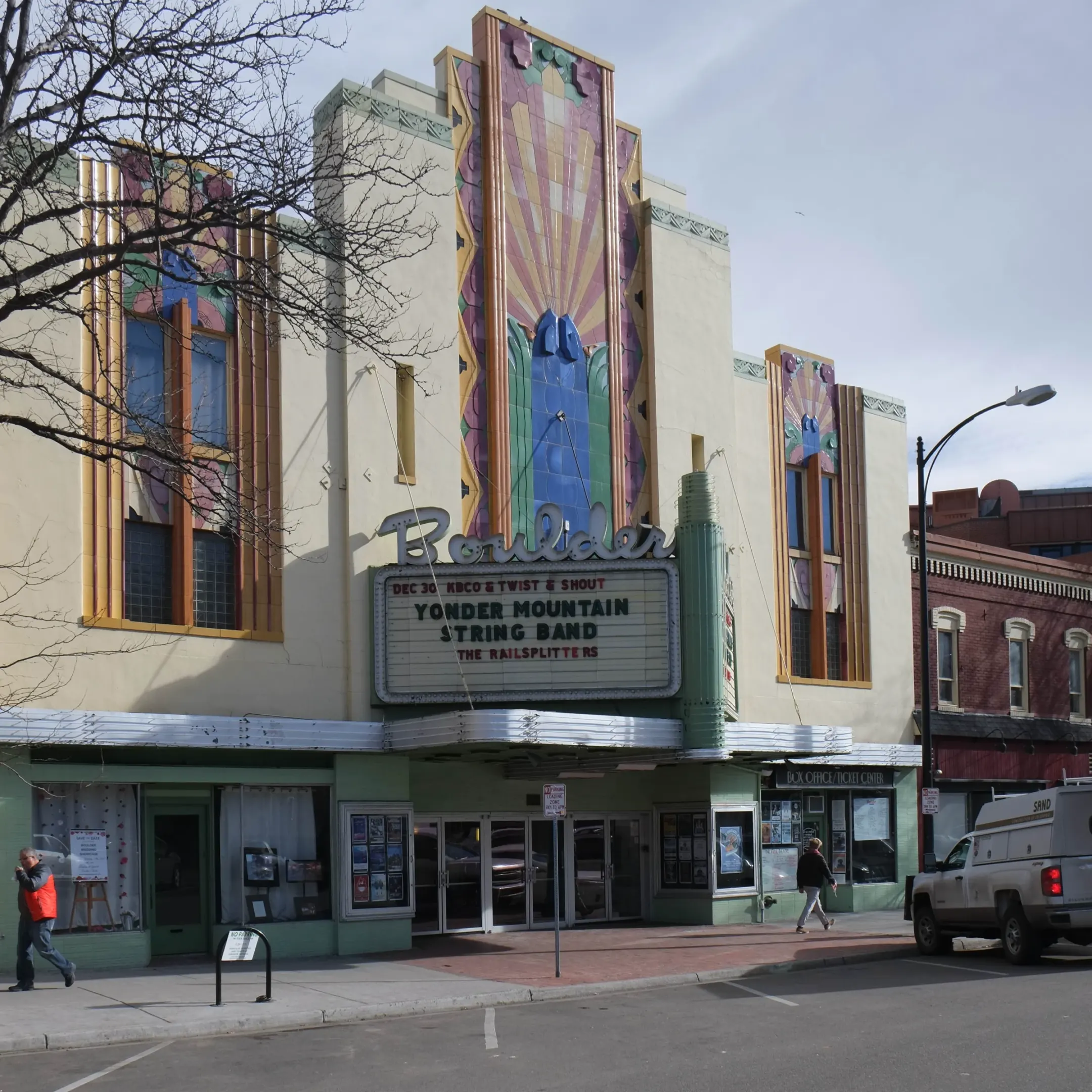 Cream stucco Art Deco theater front with tall pastel terracotta sunburst panels above a Boulder marquee and silver canopy.