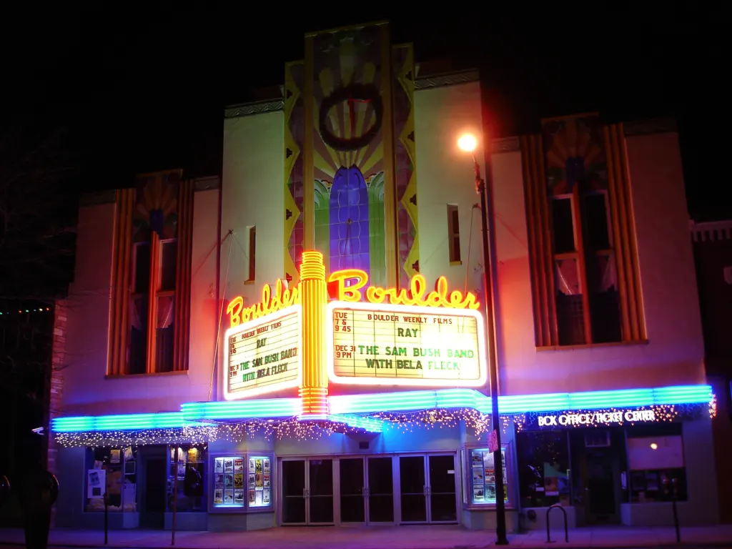 Art Deco theater facade at night with glowing orange neon Boulder signs, a lit two-sided marquee and blue neon canopy bands.
