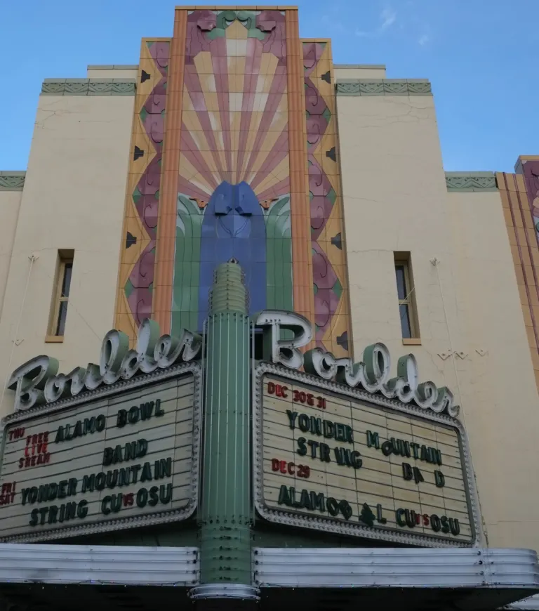 Low-angle view of a pink, yellow and blue terracotta sunburst tower rising above a two-sided Boulder marquee under blue sky.
