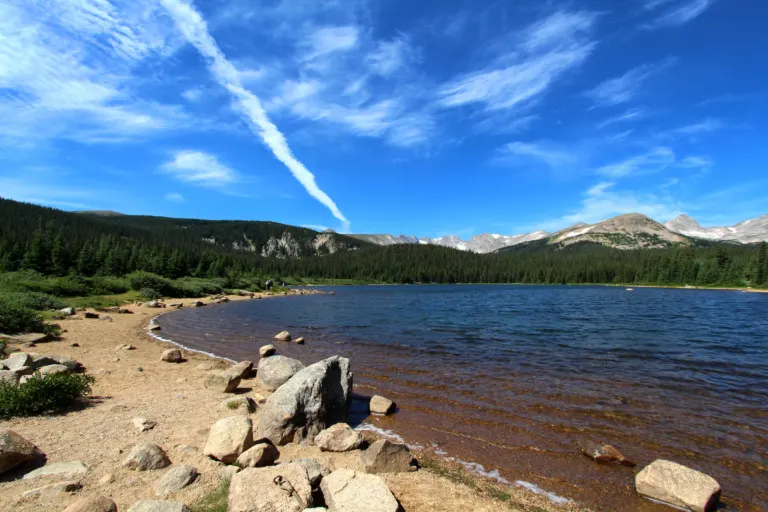 Boulders and a sandy beach along a blue mountain lake, with spruce forest and distant rocky peaks under a streaked blue sky.