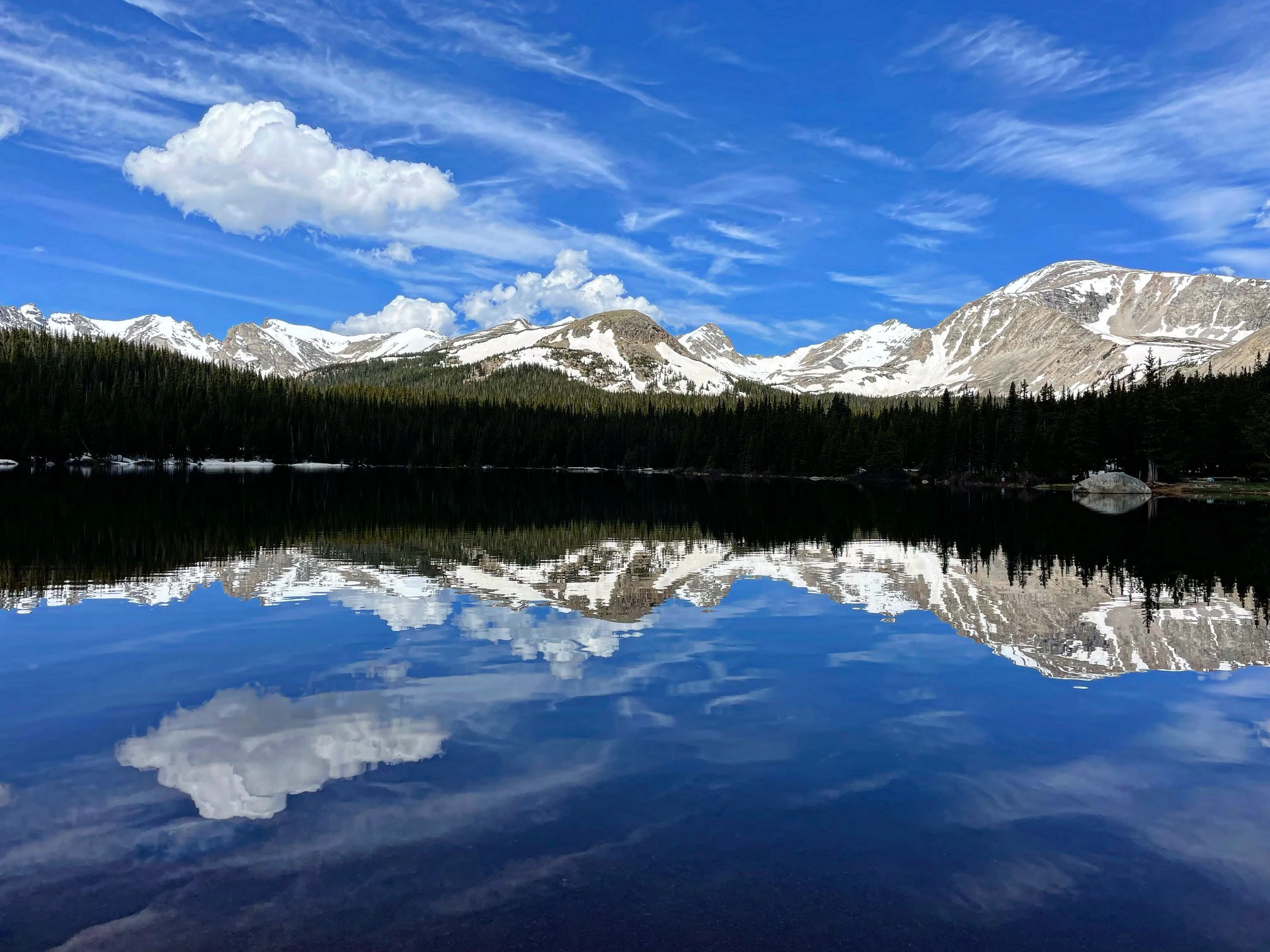 Snow-covered peaks and dark conifer forest mirrored in a still blue mountain lake under wispy clouds.