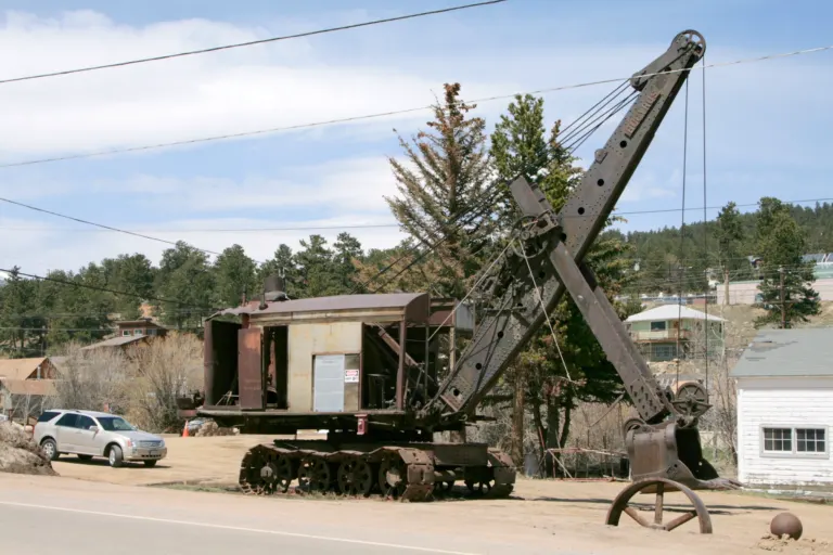 Rusted steam shovel on crawler tracks with its long boom raised, beside a white shed, pine trees and a parked SUV.
