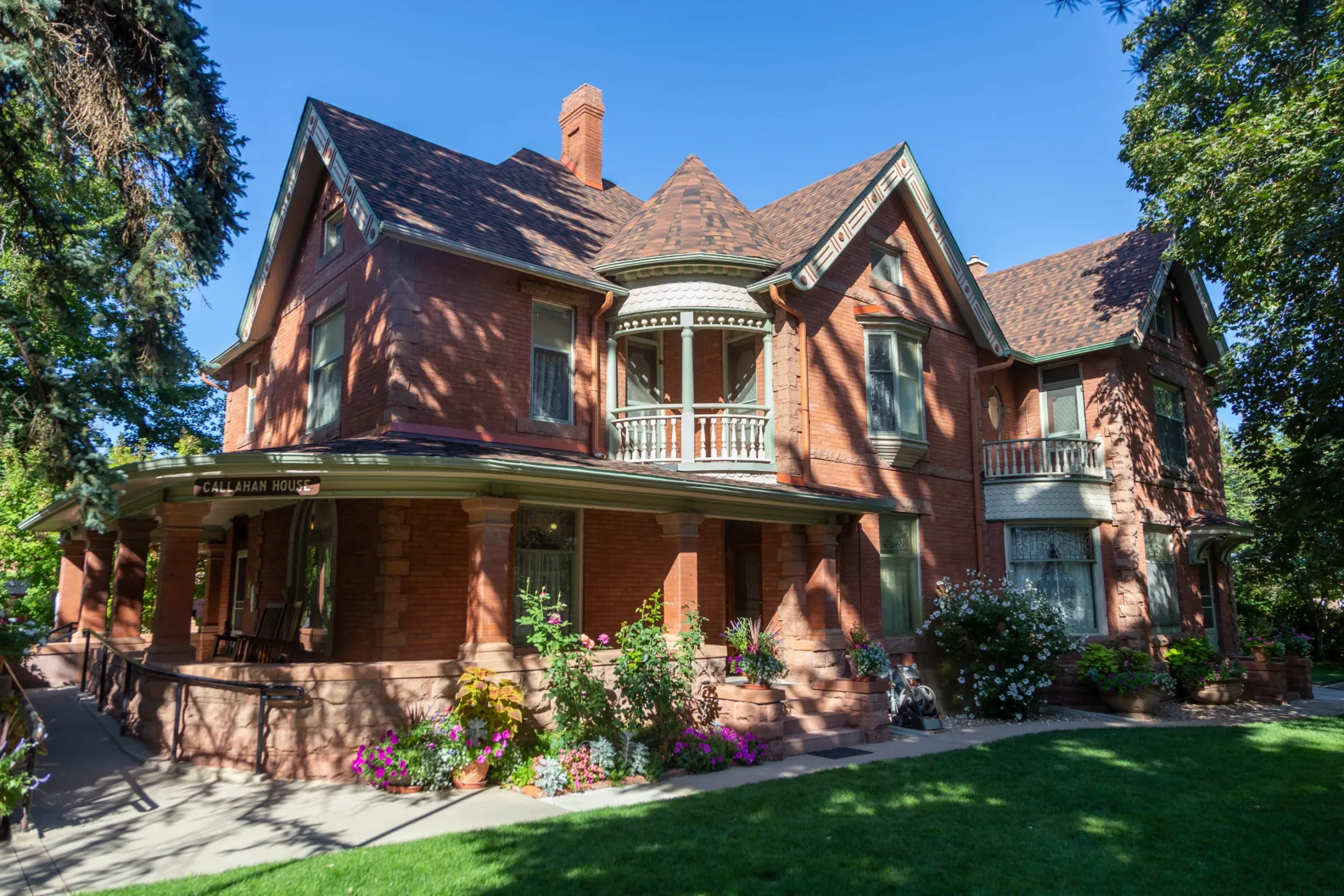 Red brick Queen Anne house with a curved wraparound porch, a round turret balcony and steep gables, seen across a lawn under blue sky.