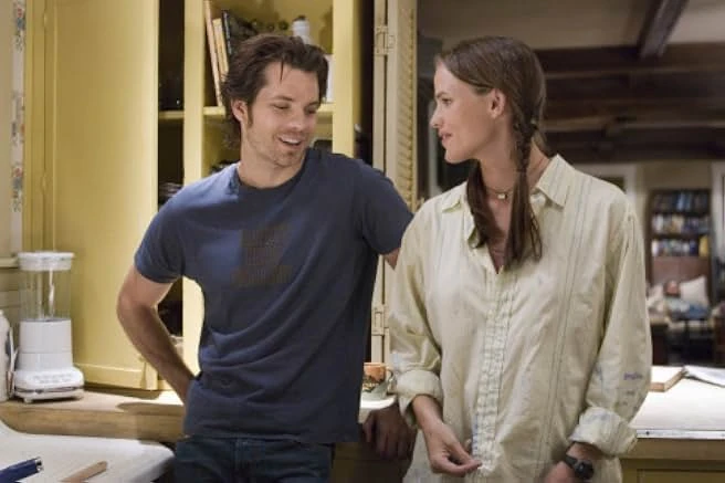 A smiling man in a navy T-shirt leans toward a woman with a long braid and a cream shirt in a kitchen.