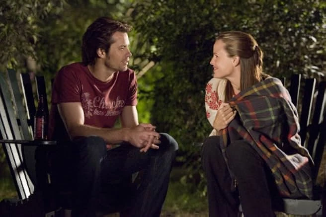 A man in a red T-shirt and a woman wrapped in a plaid blanket smile at each other from wooden chairs at night.