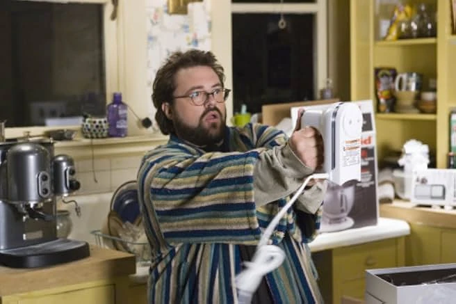 A bearded man in glasses and a striped bathrobe aims a white kitchen mixer across a yellow kitchen.