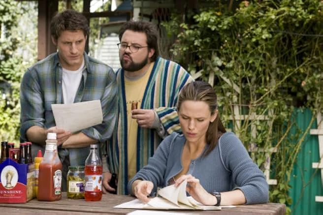 A woman reads papers at an outdoor table of condiments while a man in plaid and a bearded man in a striped robe look on.