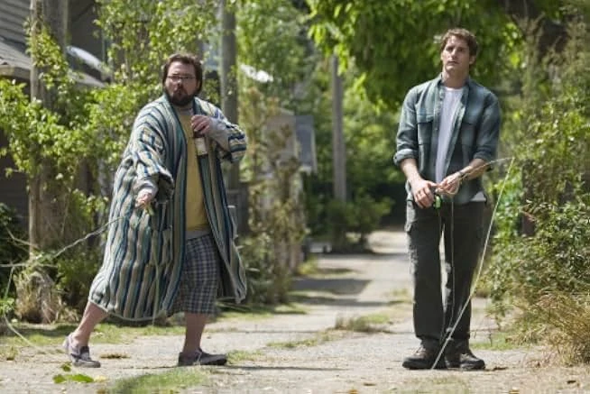 A bearded man in a striped bathrobe and a man in a plaid shirt hold taut strings in a leafy gravel alley.