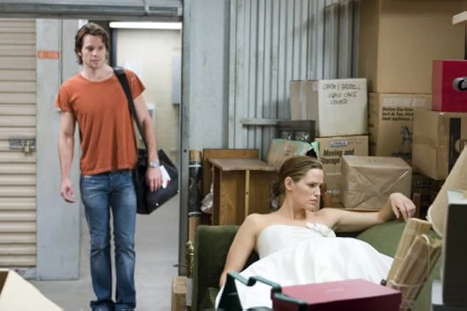 A woman in a strapless white wedding dress lounges among stacked boxes in a storage unit as a man in an orange T-shirt walks in.